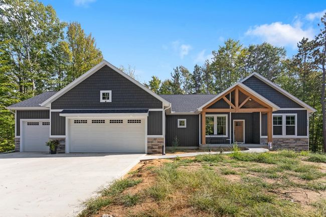 A modern gray house with a two-car garage, stone veneer base, and wood-framed entrance, set against a wooded backdrop.