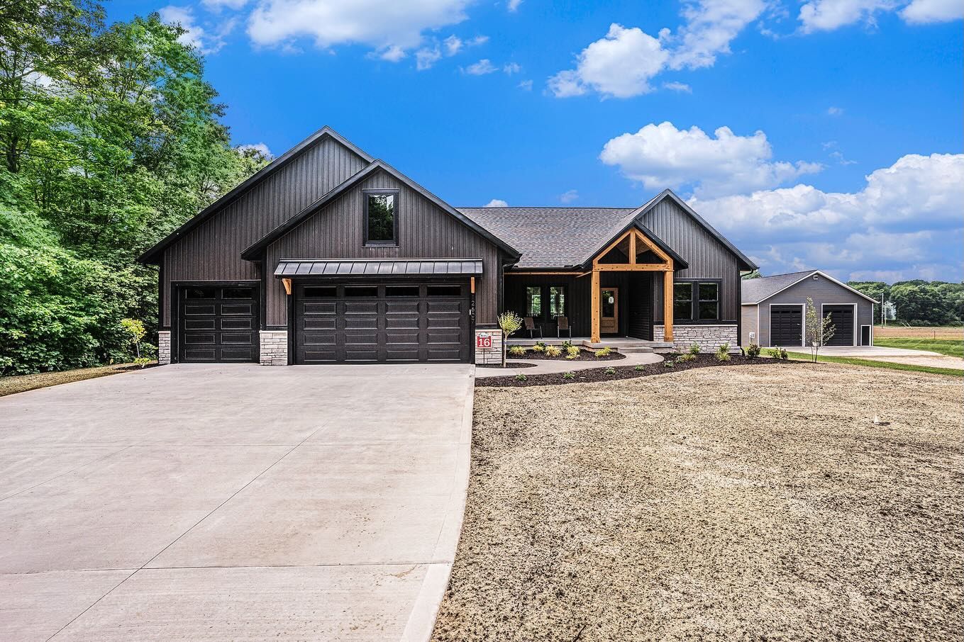 A modern farmhouse with dark vertical siding, stone accents, a multi-car garage, and a wood-framed porch under a blue sky.