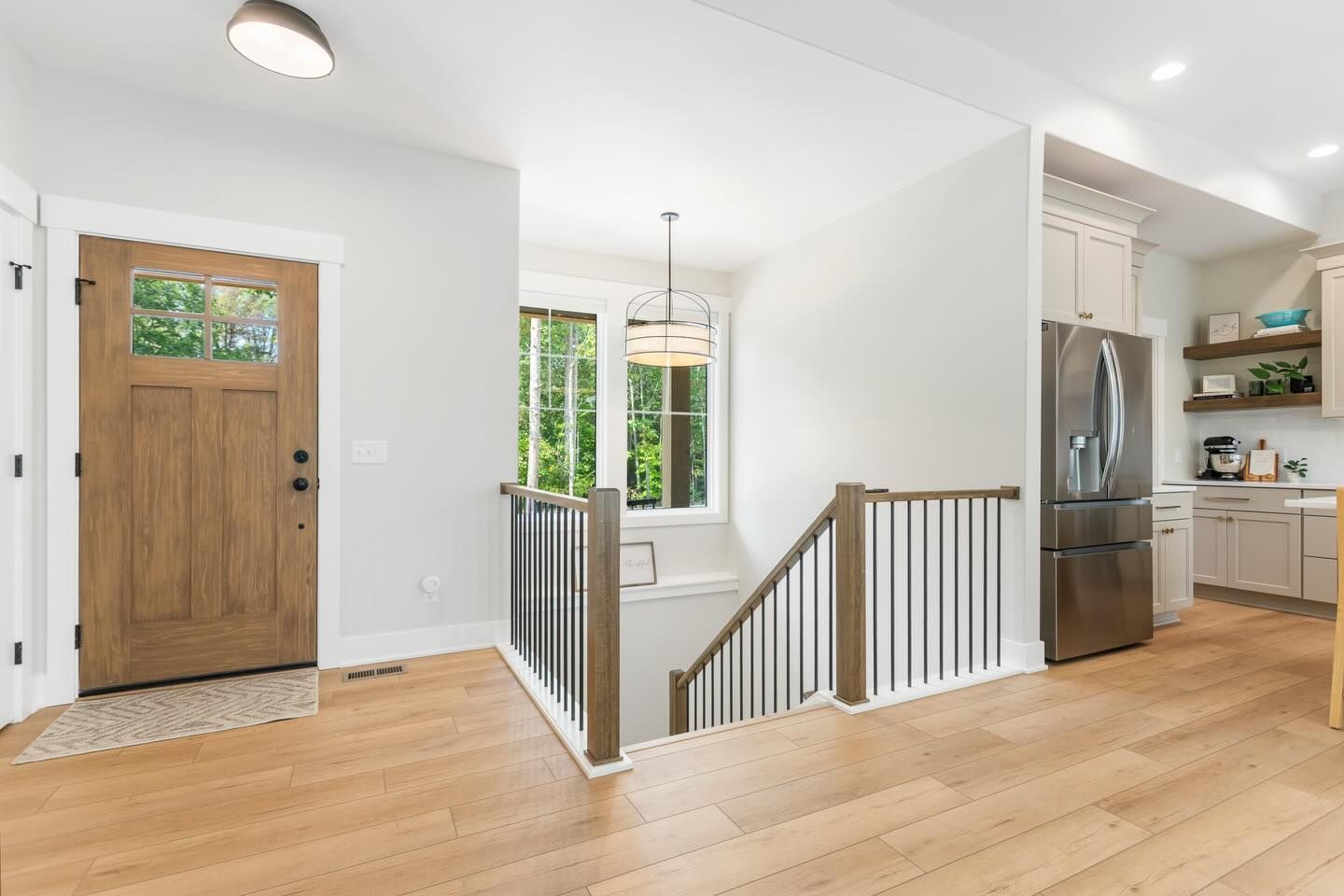 An entryway with a wooden door, hardwood floors, a staircase with metal railings, and a view into a bright kitchen.