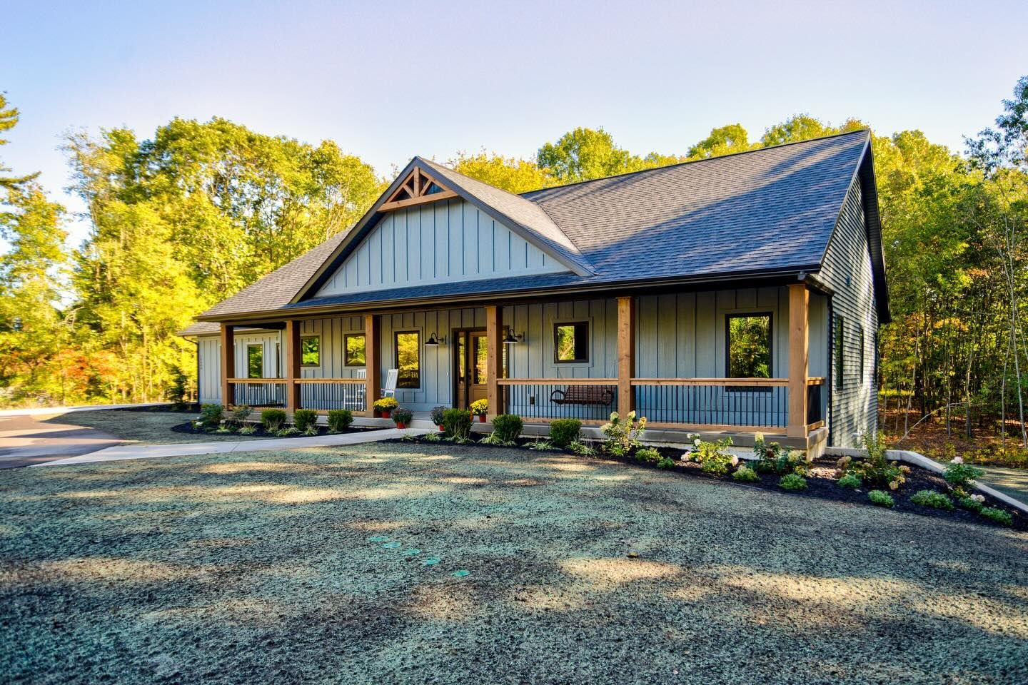 A modern gray farmhouse with a large front porch and stone siding, surrounded by trees on a sunny day.