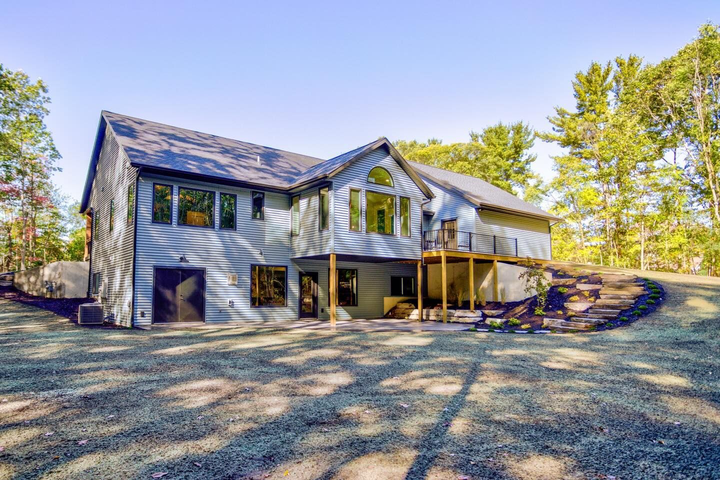 A large, two-story house with gray horizontal siding, dark accents, and a back deck, set in a wooded area with gravel ground.