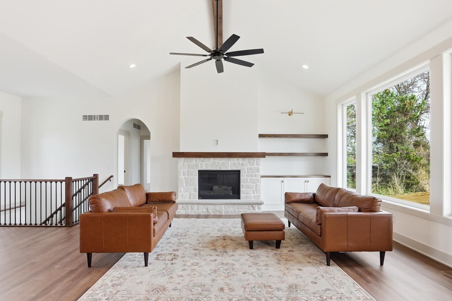 A spacious, bright living room features two tan leather sofas, a central ottoman, a stone fireplace, and a ceiling fan.