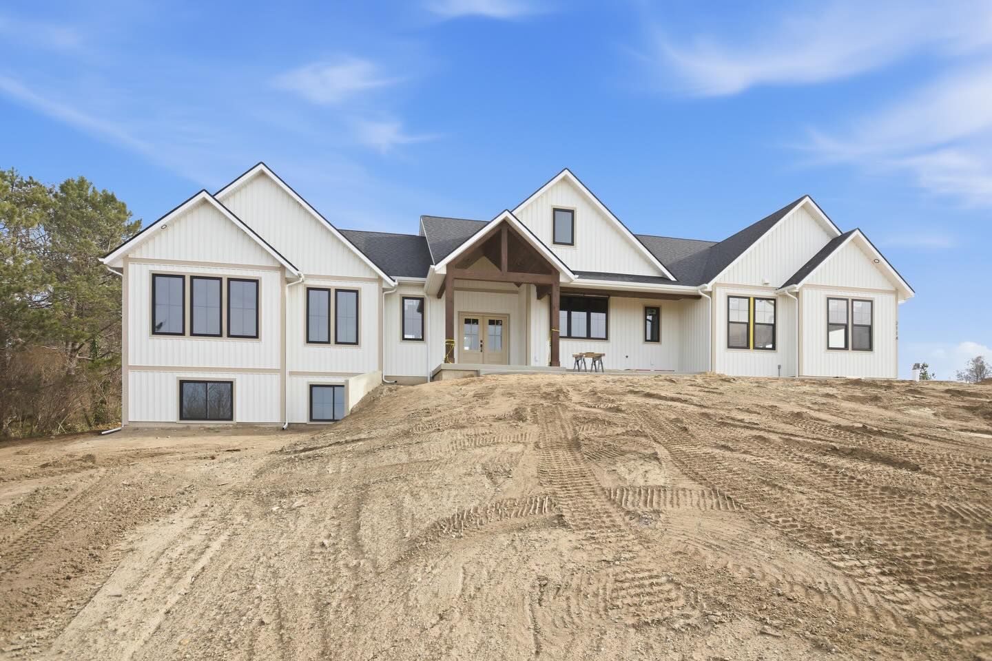 Newly built white farmhouse with a dark roof on a dirt lot under a bright blue sky.