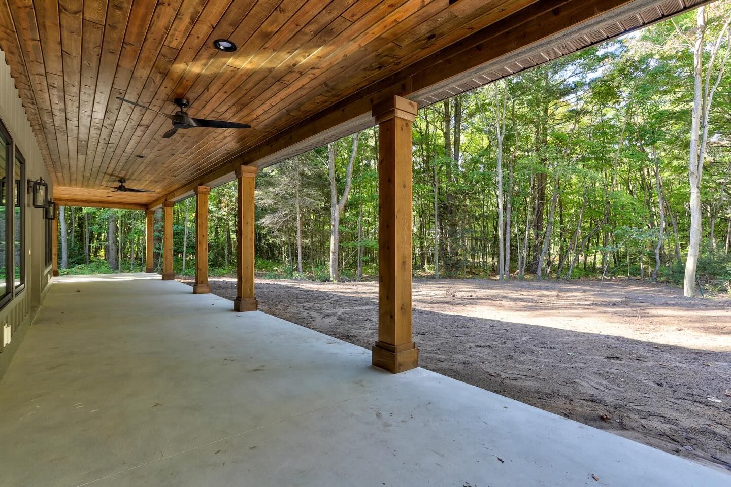 A covered patio with a wooden ceiling and ceiling fans, overlooking a wooded yard with exposed dirt.