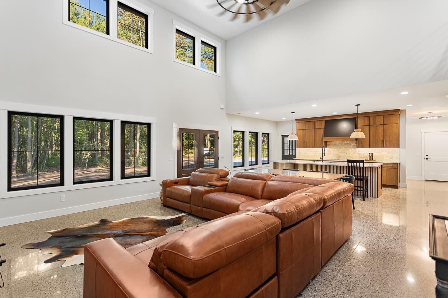 Spacious living area with a large leather sectional, high ceilings, tall windows, and an open kitchen with wooden cabinets.