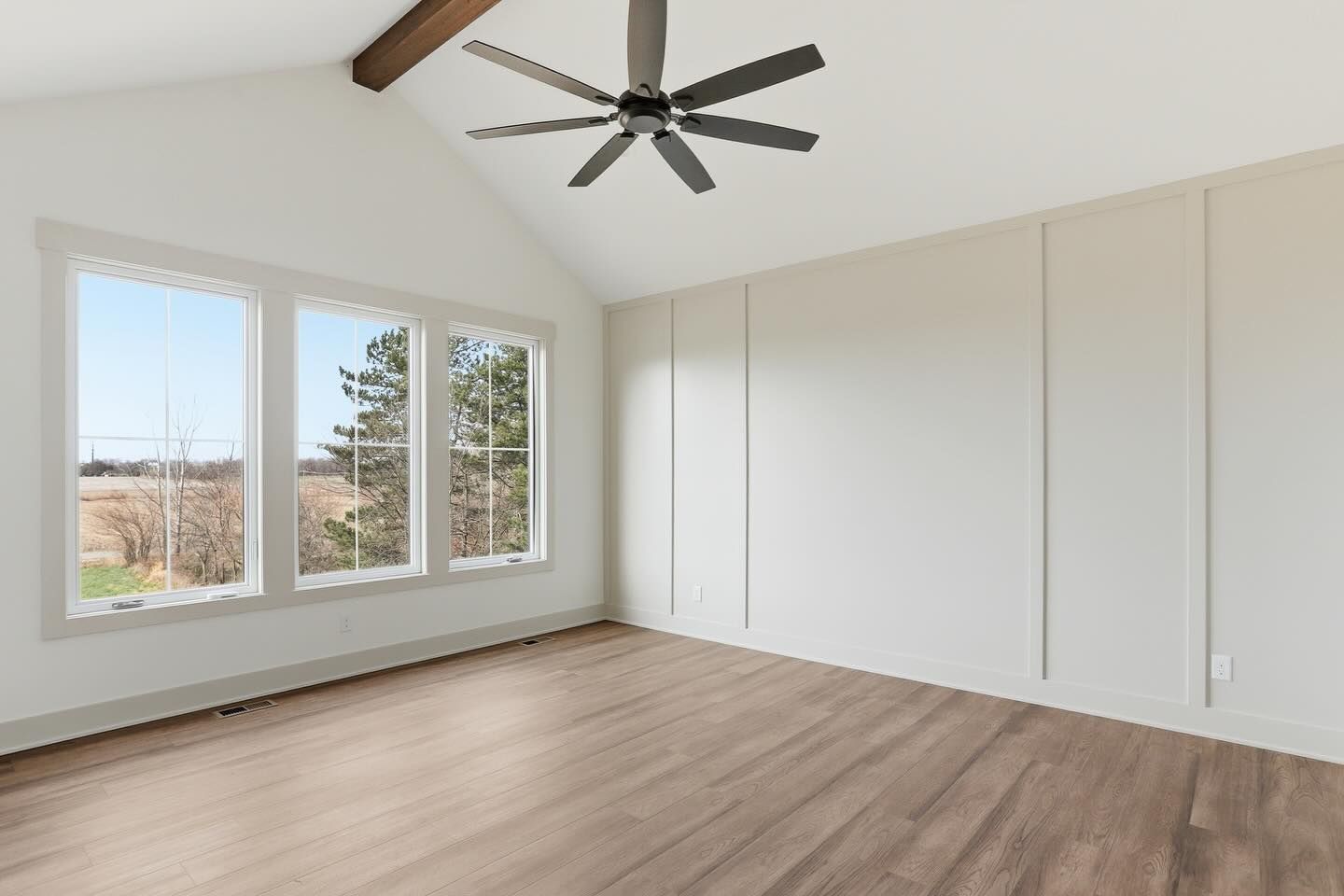 A light-filled, empty room with wood floors, vaulted ceilings, a ceiling fan, and white paneled walls.