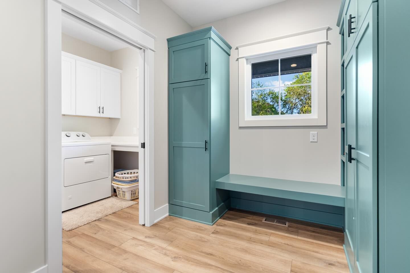 A mudroom with teal cabinets and a bench, looking into a laundry room featuring a white washer, dryer, and cabinetry.