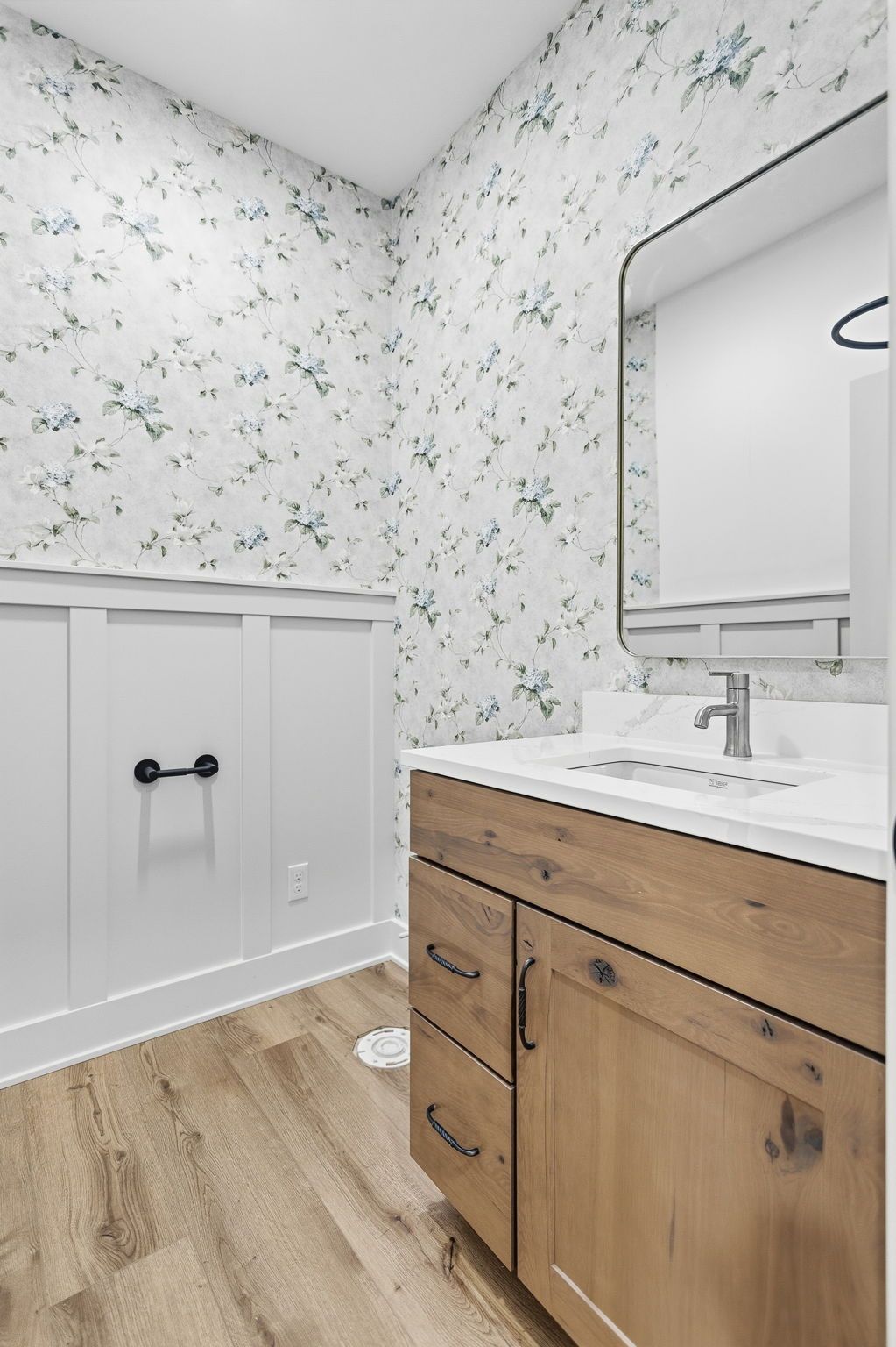 Modern bathroom with floral wallpaper, wood vanity, white sink, mirror, and wainscoting.