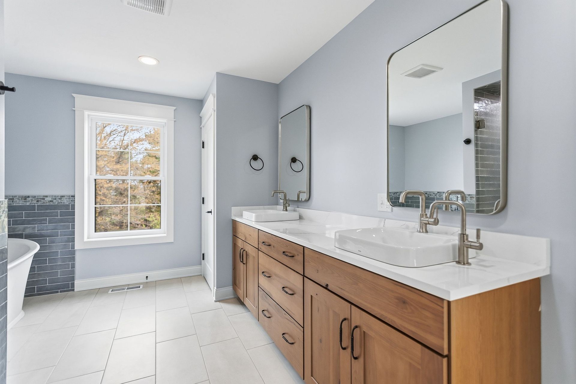 Bright bathroom with double wood vanity, white countertops, mirrors, and a windowed shower area