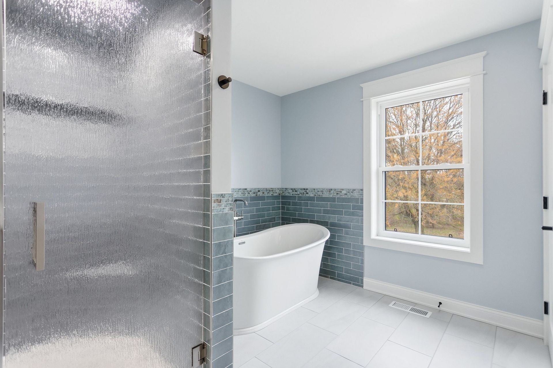 Modern bathroom with frosted glass shower door, freestanding tub, blue tile accent wall, and window.