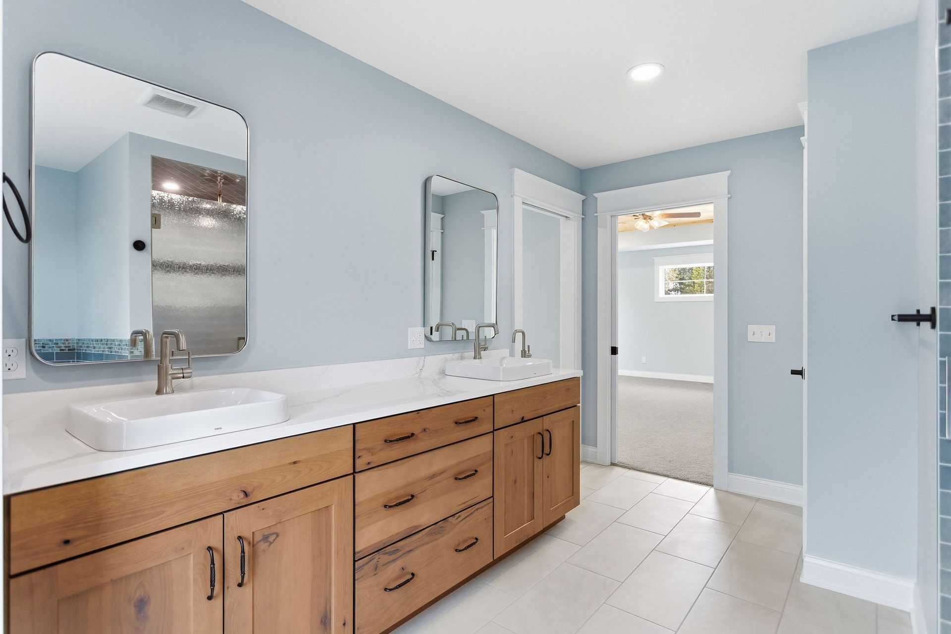 Bright bathroom with wood vanity, white countertop, mirrors, and open doorway to a shower area
