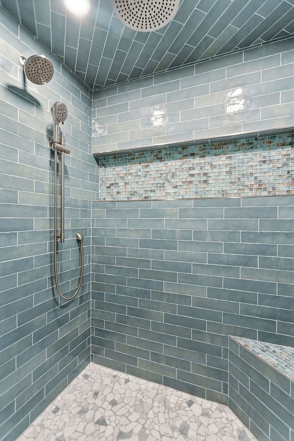 Blue-tiled walk-in shower with rainfall head, handheld sprayer, mosaic accent strip, and bench seat