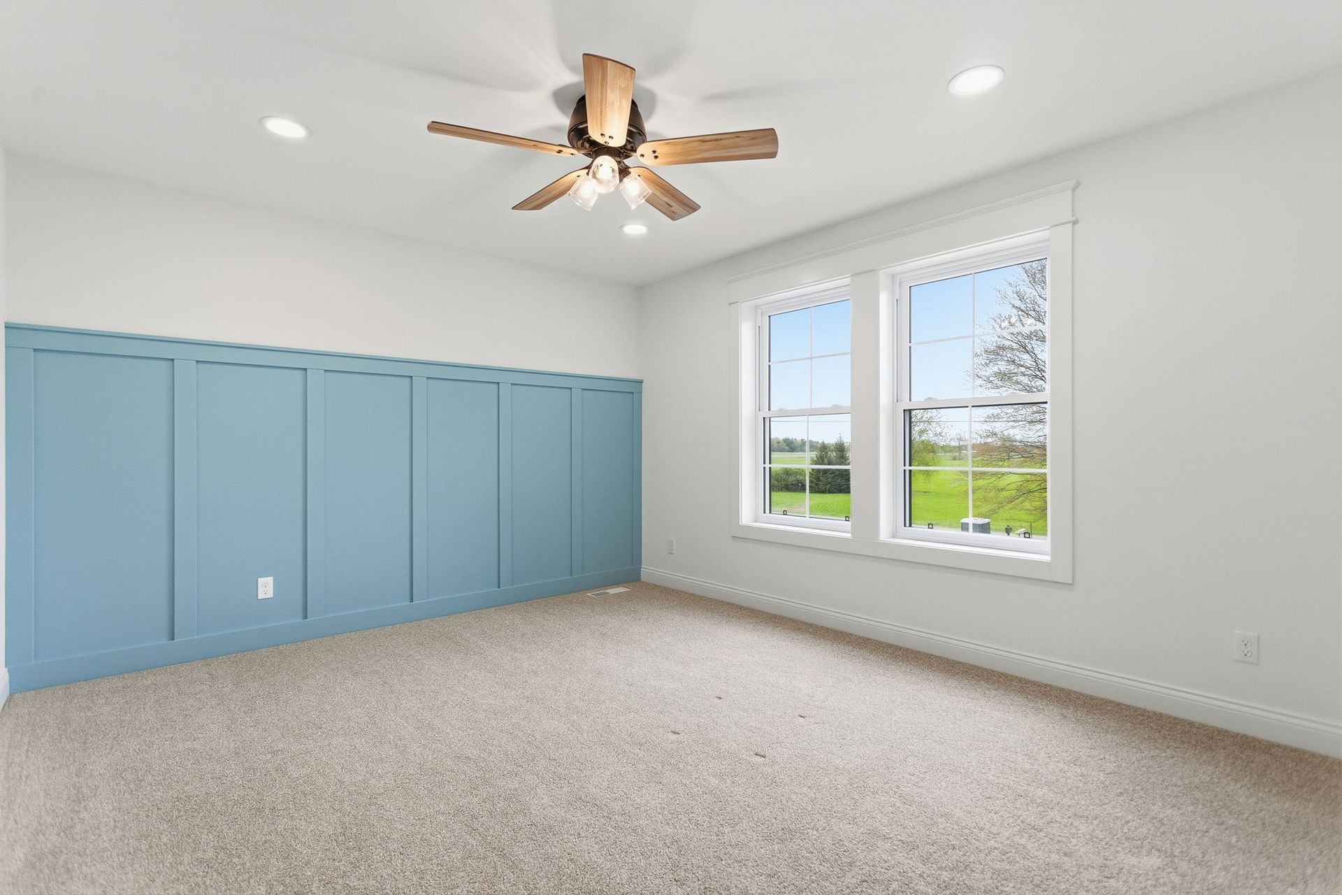 Empty bedroom with blue built-in cabinets, beige carpet, ceiling fan, and two windows.