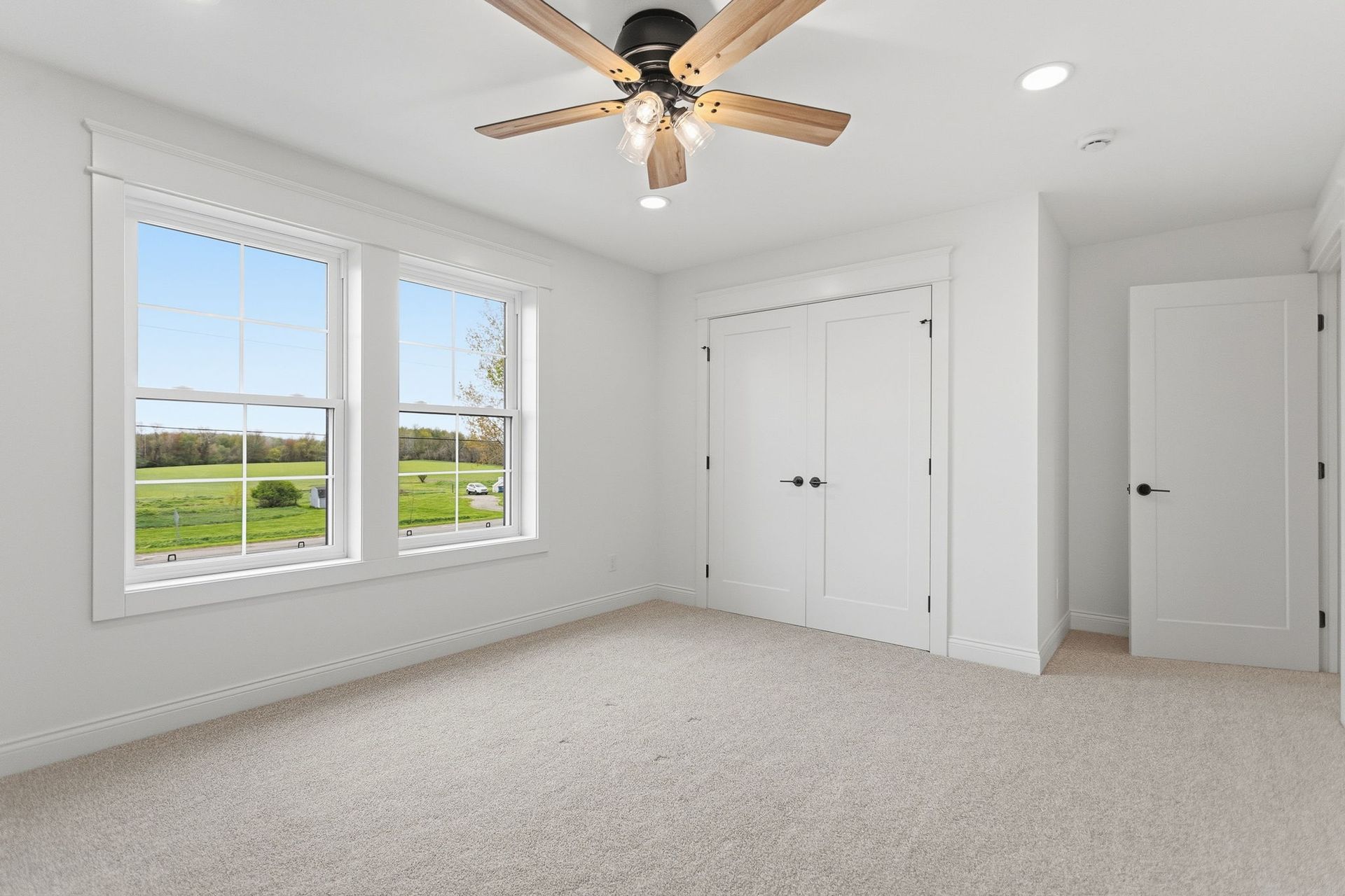 Bright empty bedroom with carpet, ceiling fan, white double closets, and large windows overlooking a green field