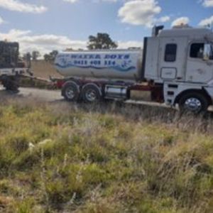 A white water tanker truck parked on a dirt roadside next to a field of dry, tall grass under a blue, cloudy sky.