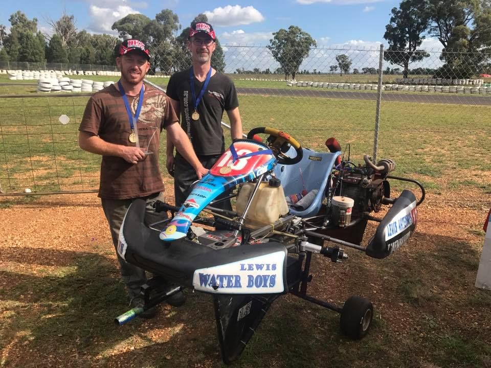 Two people wearing medals stand beside a racing go-kart on a dirt track, with 