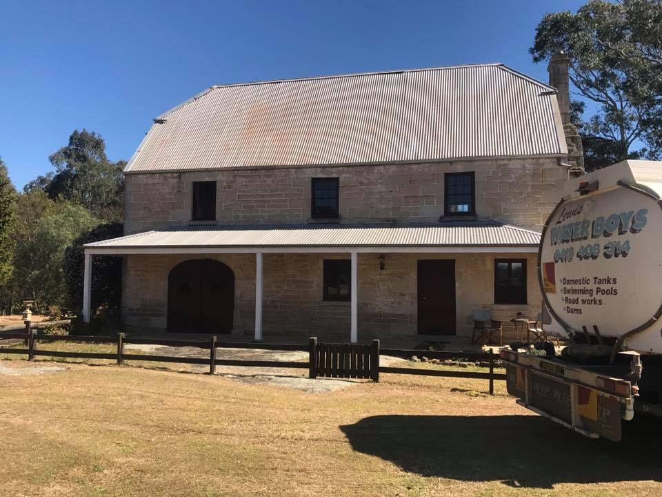 A historic two-story stone house with a metal roof and front veranda, next to a parked water tanker truck.