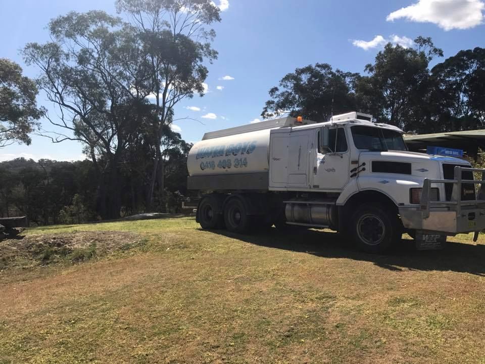A white tanker truck parked on a grass field near trees under a blue, partly cloudy sky.