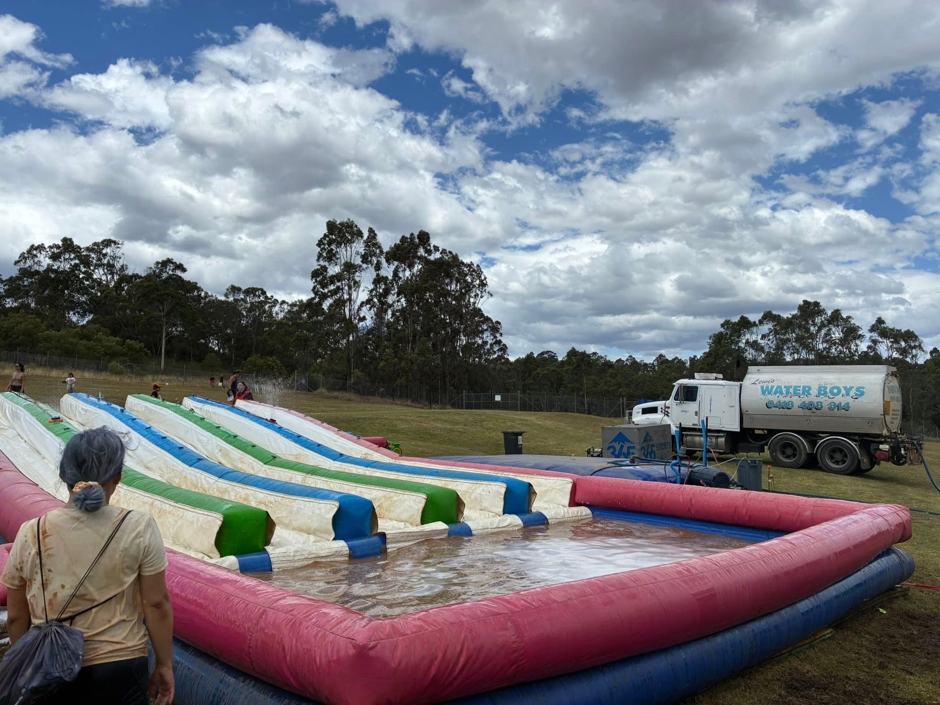 A person stands before a large, multi-lane inflatable water slide in a grassy field with a water truck parked nearby.