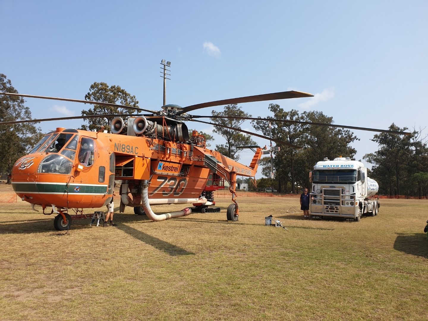 A large orange helicopter parked on a grassy field next to a white fuel tanker truck under a clear blue sky.