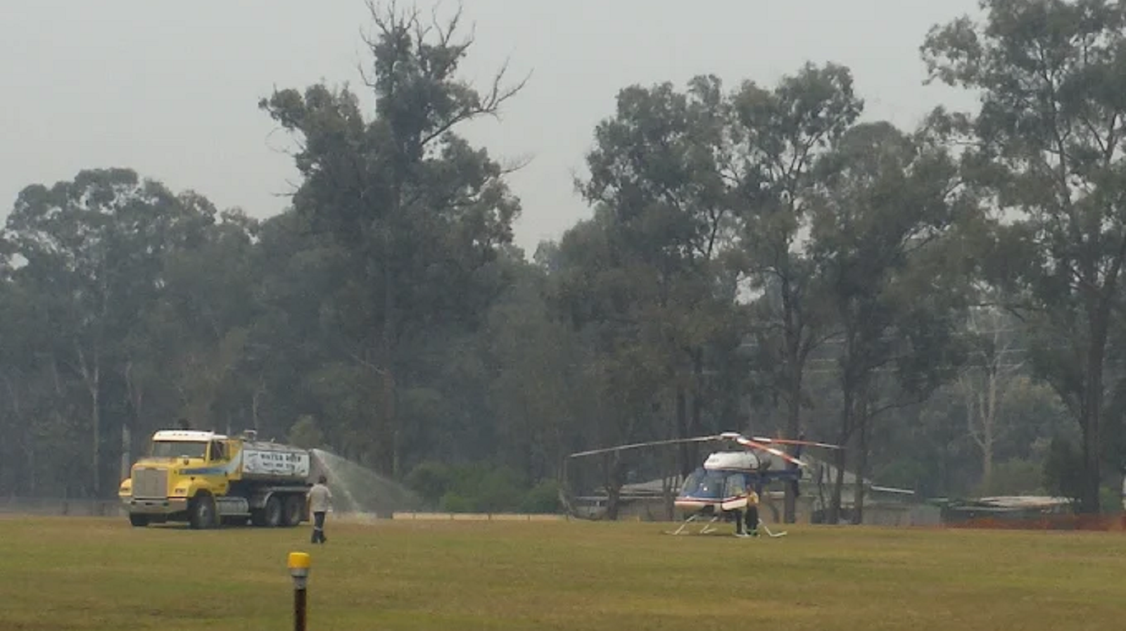 A yellow tanker truck sprays water near a helicopter on a grassy field with trees in the background under a hazy sky.