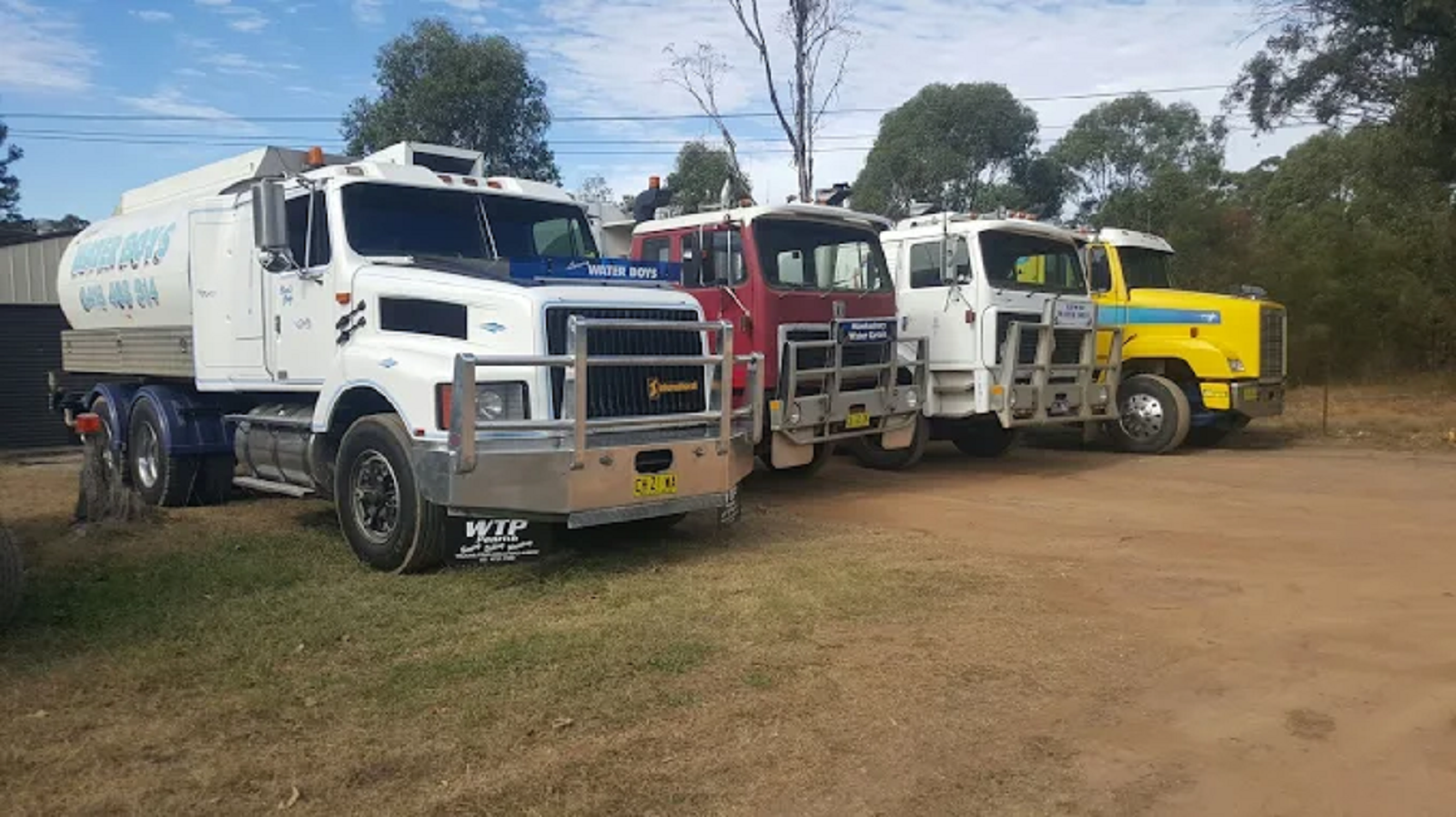 A line of four work trucks, colored white, red, white, and yellow, parked on a dirt lot with trees in the background.