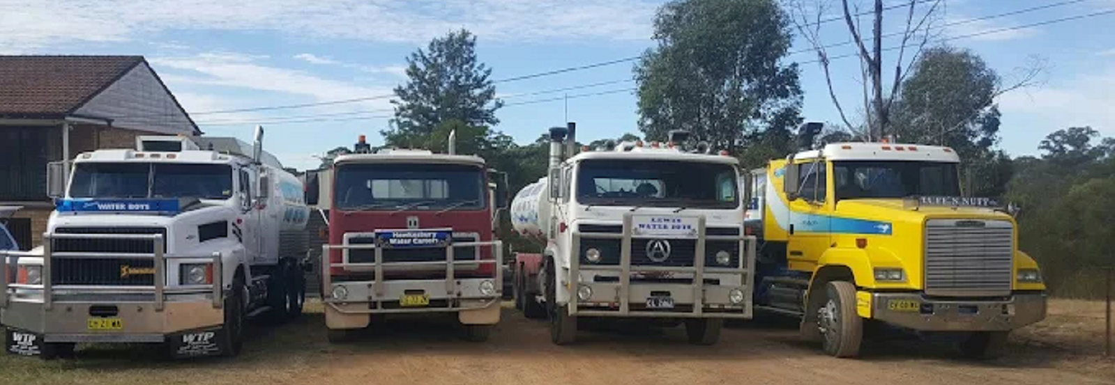 Four tanker trucks, colored white, red, white, and yellow, parked side-by-side in a dirt lot under a bright sky.