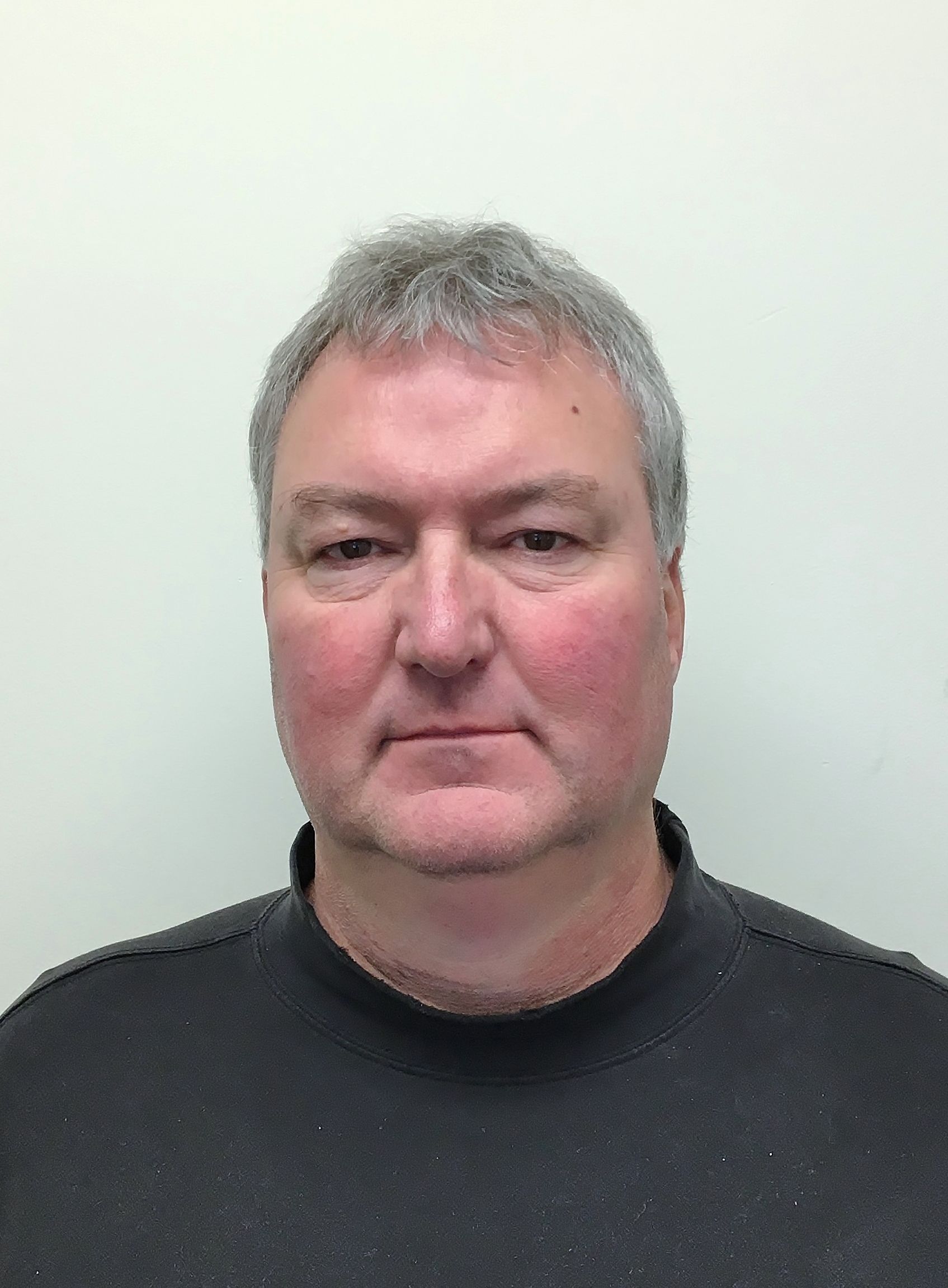 A man in a black sweater is standing in front of a white wall.