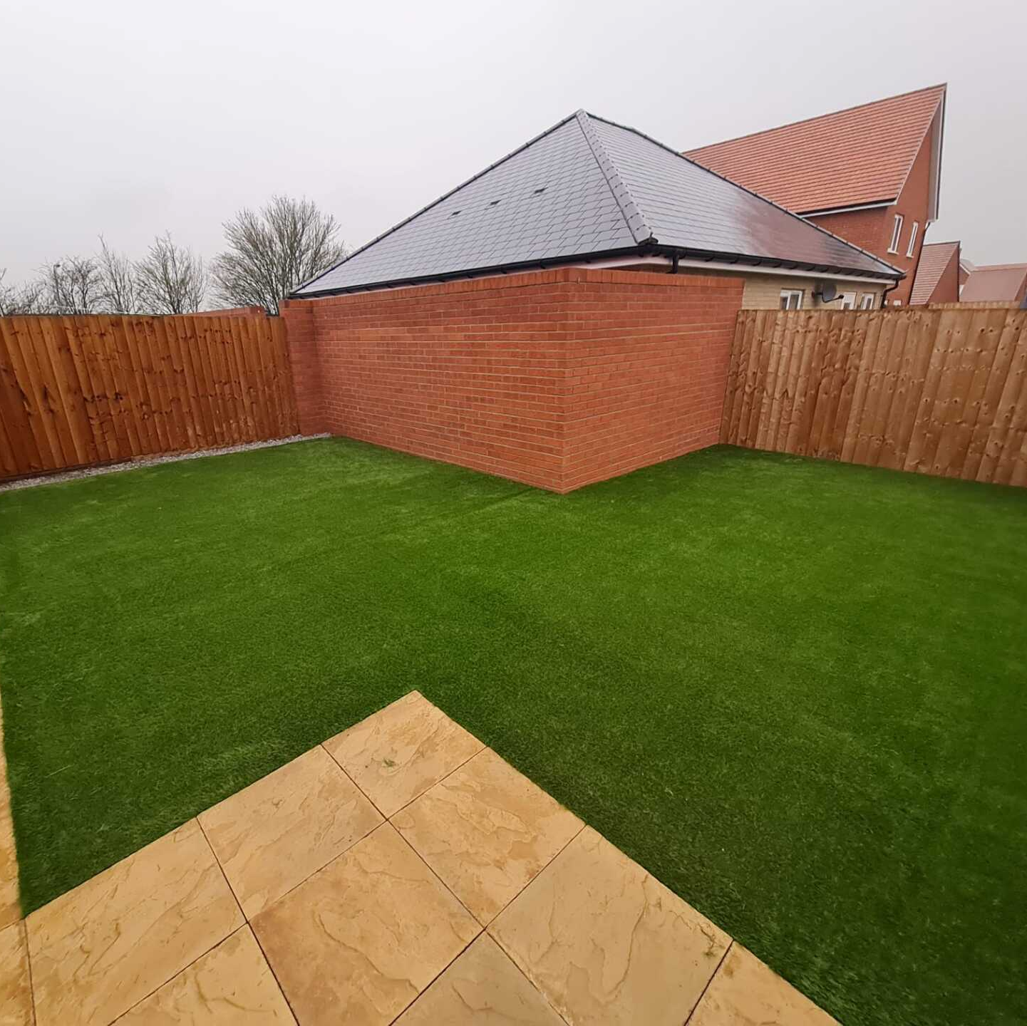 A backyard with a brick wall , a wooden fence , and a lush green artificial grass lawn.