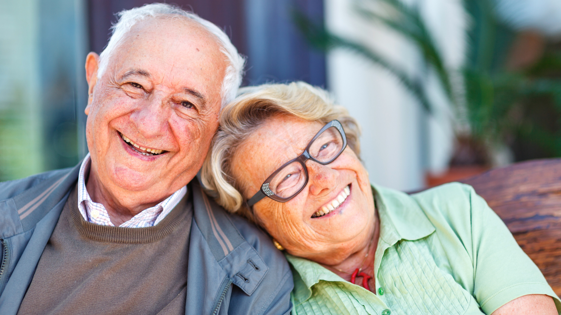 Smiling elderly couple together outdoors; woman rests head on man's shoulder.