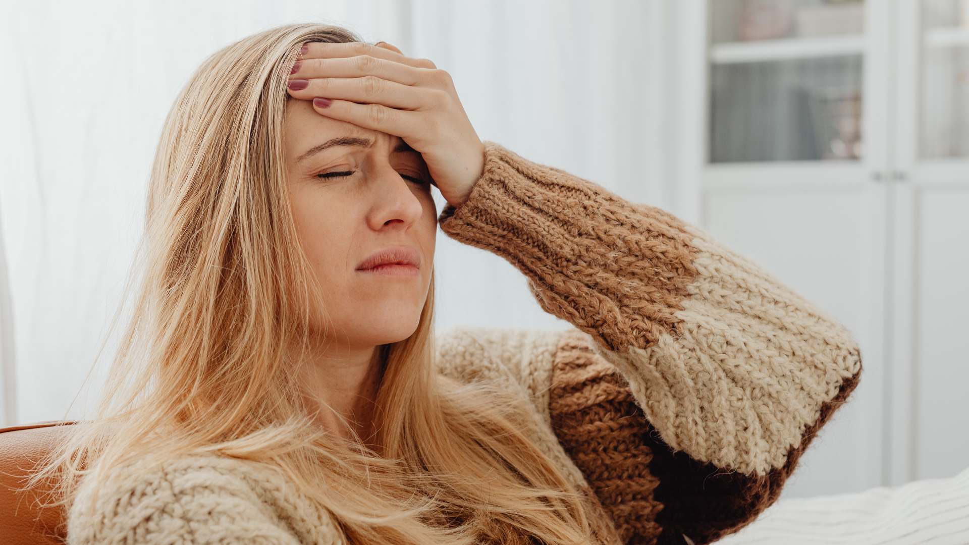 Woman with long blonde hair, eyes closed, hand on forehead, looking distressed, wearing a sweater.