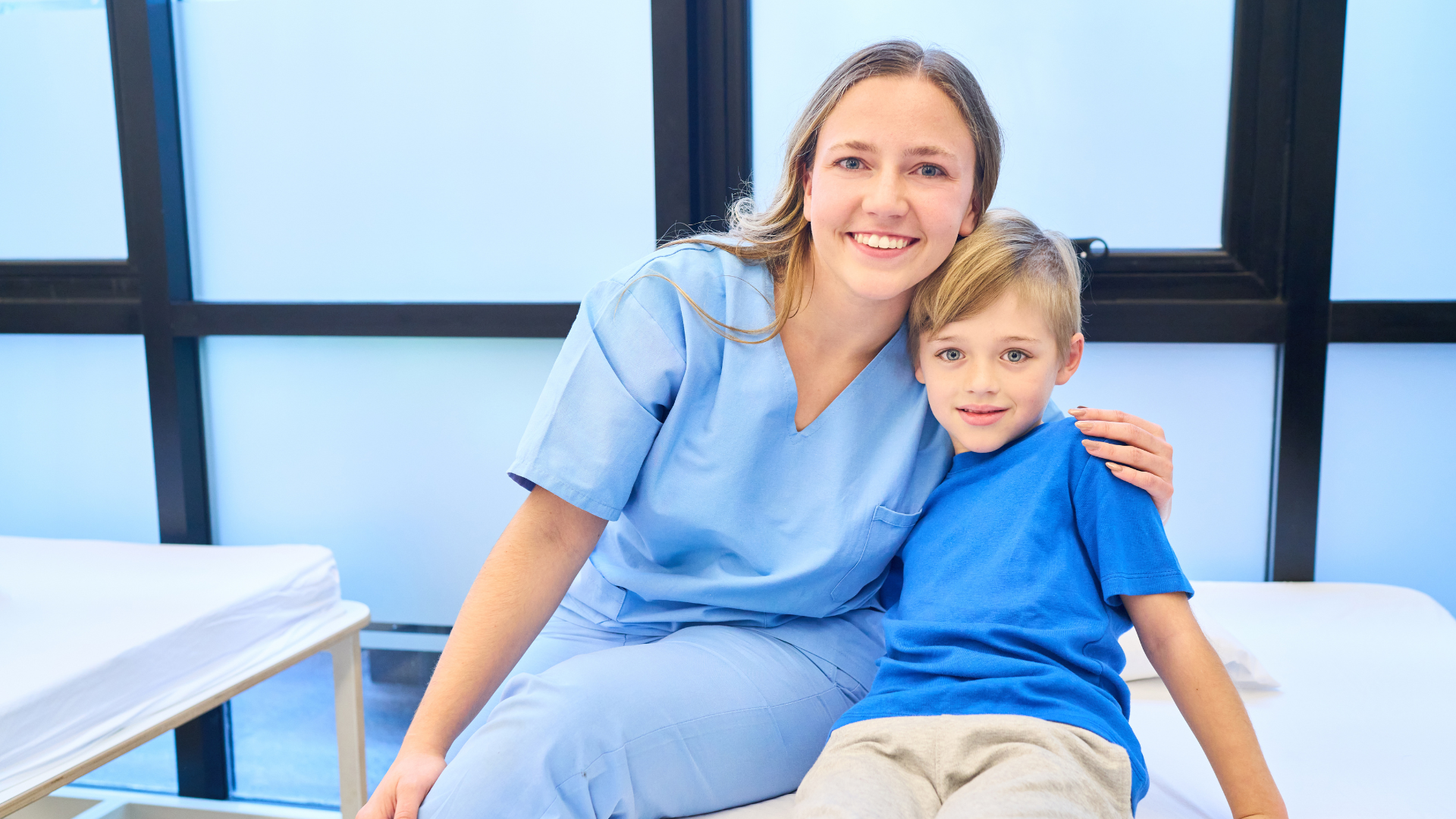 Woman in blue scrubs smiles, arm around a child in a blue shirt, seated on a medical exam table.