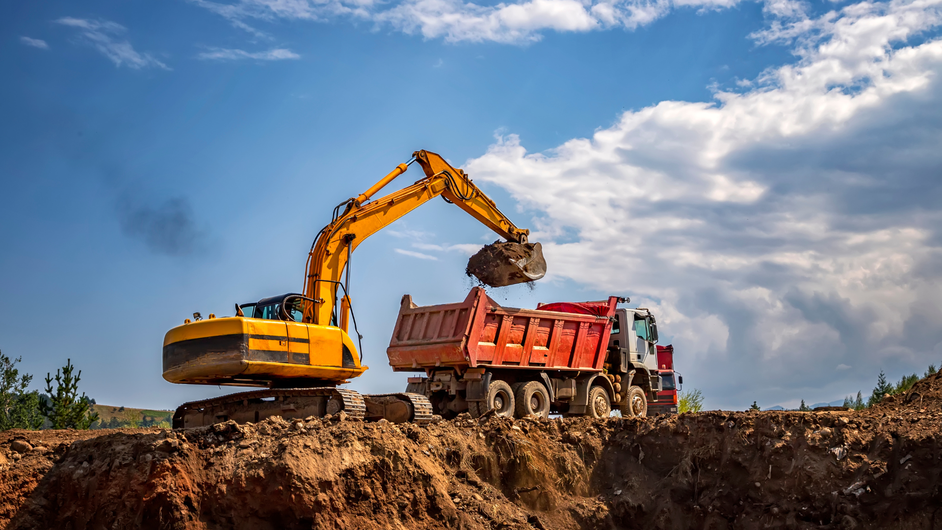 Yellow excavator loads dirt into a red dump truck on a construction site under a blue sky.