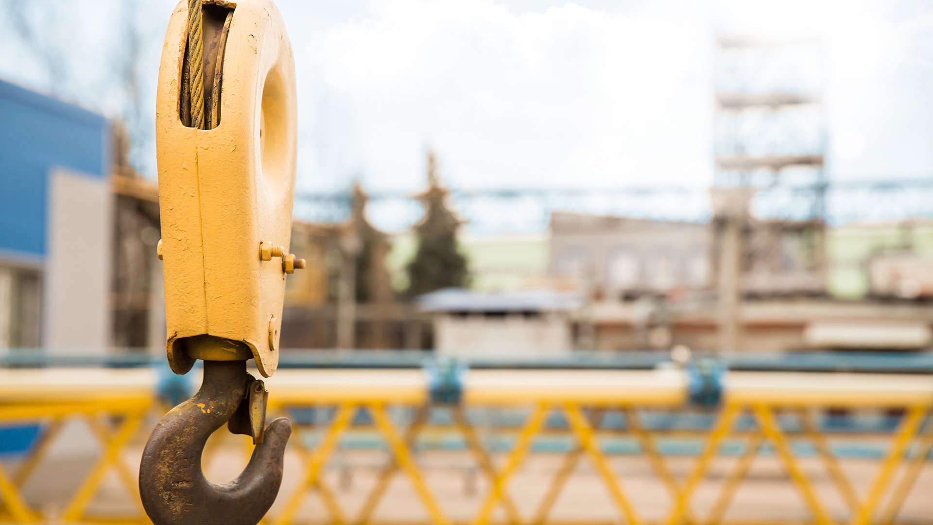 Yellow construction crane hook in foreground with blurred industrial site and safety barrier behind