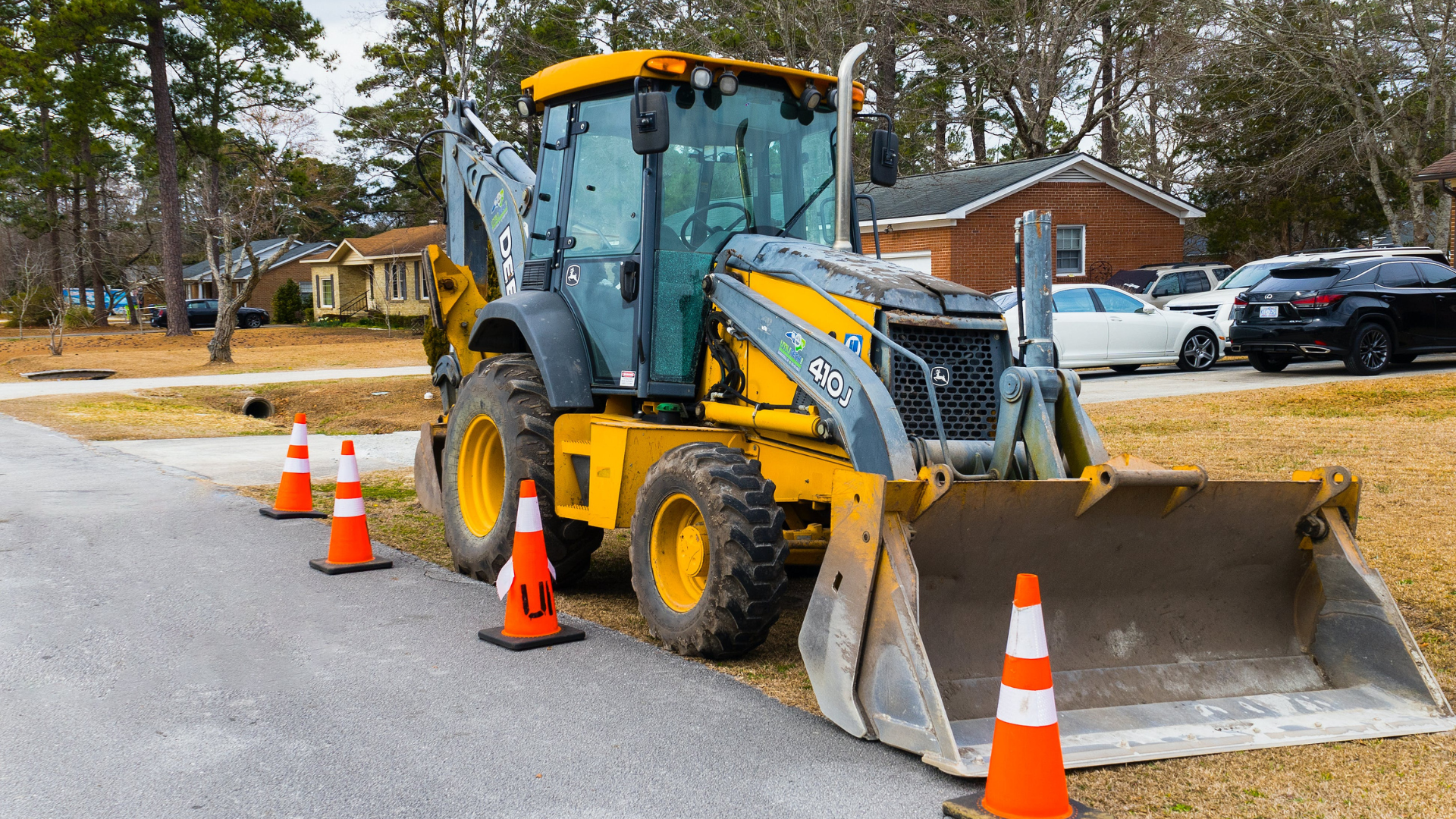 Yellow backhoe with front loader parked on a residential street beside orange traffic cones.