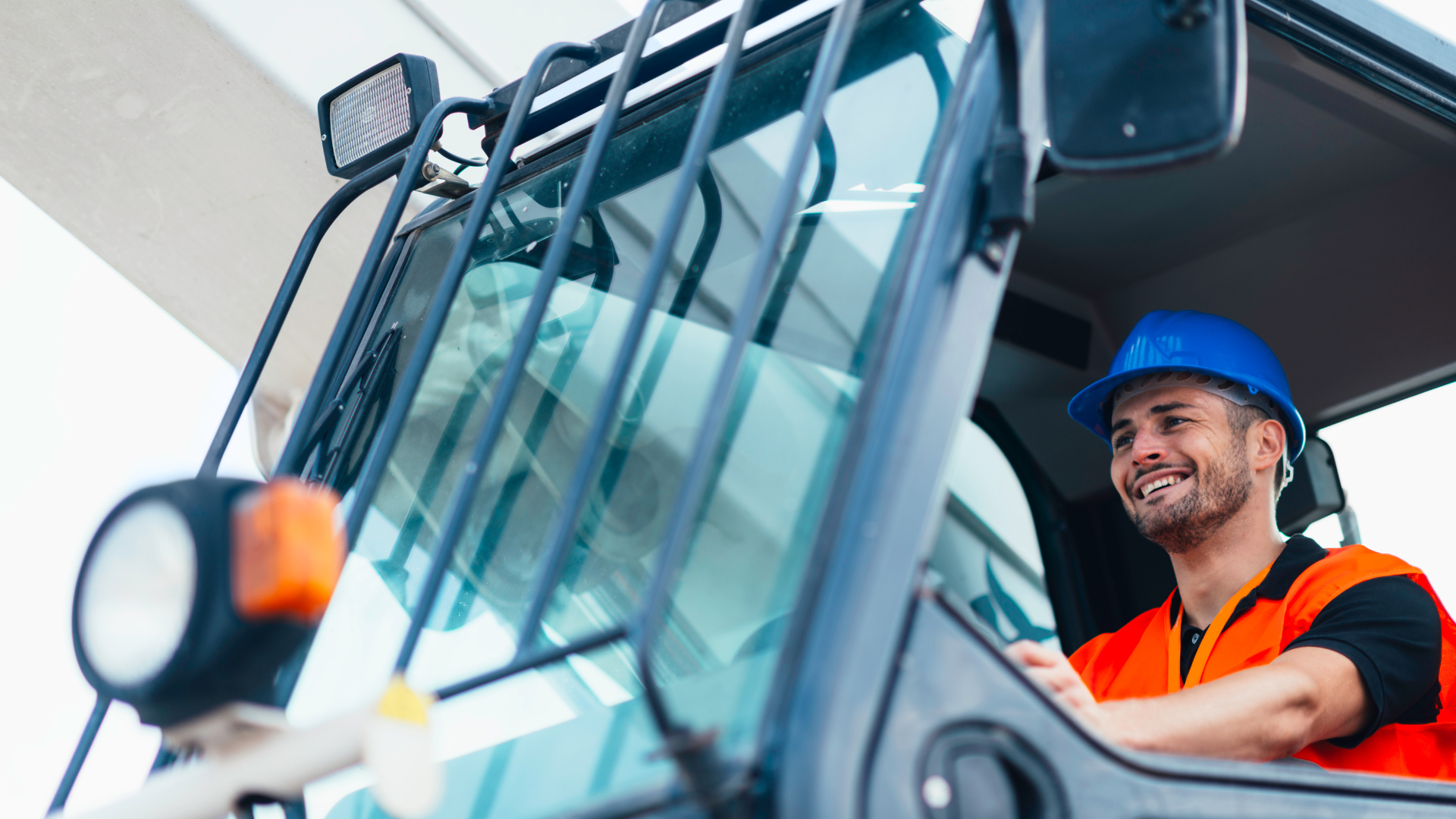 Worker in blue hard hat and orange vest operating heavy machinery, smiling in cab