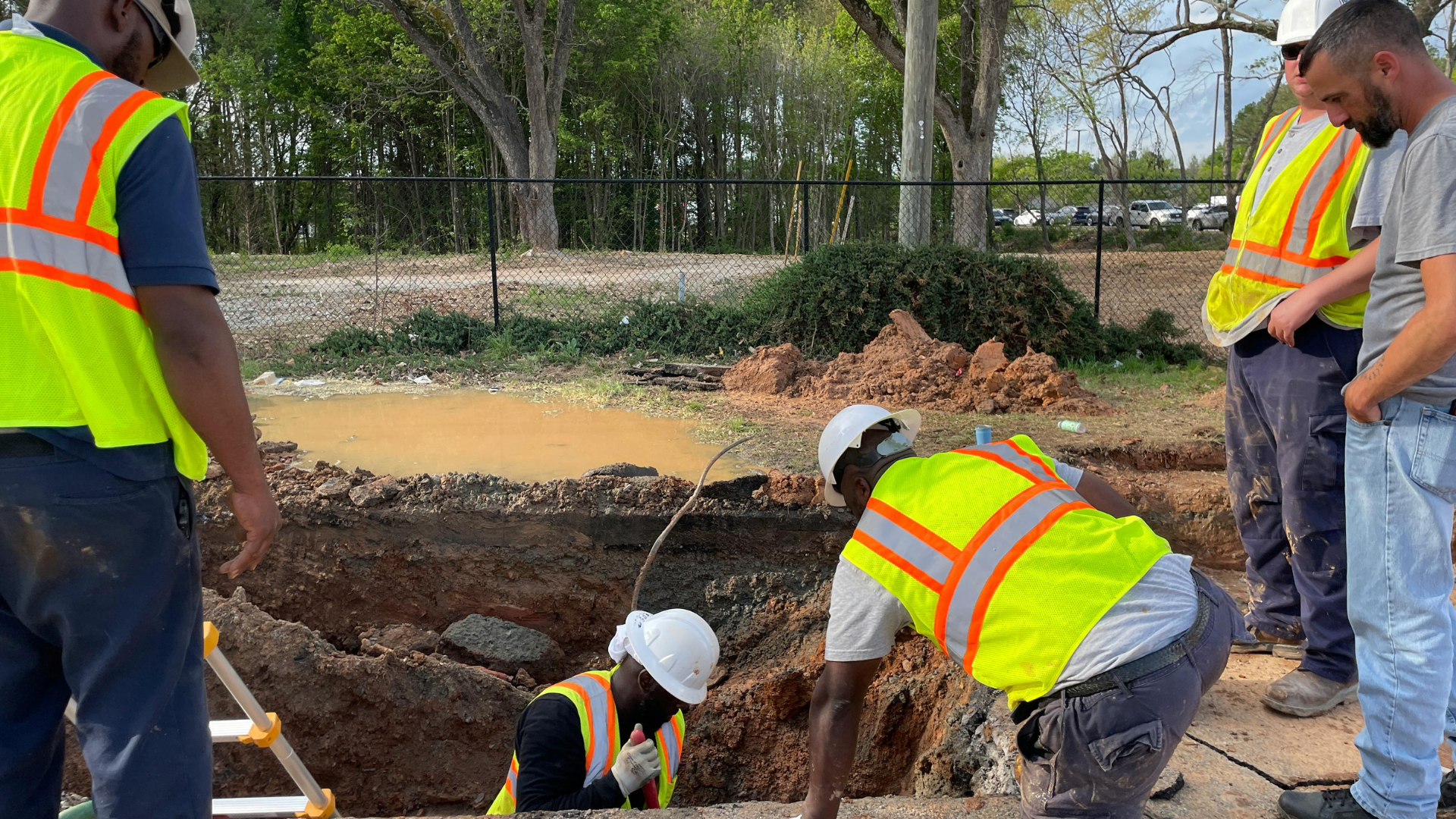 Workers in reflective vests and hard hats inspecting a muddy trench at a construction site