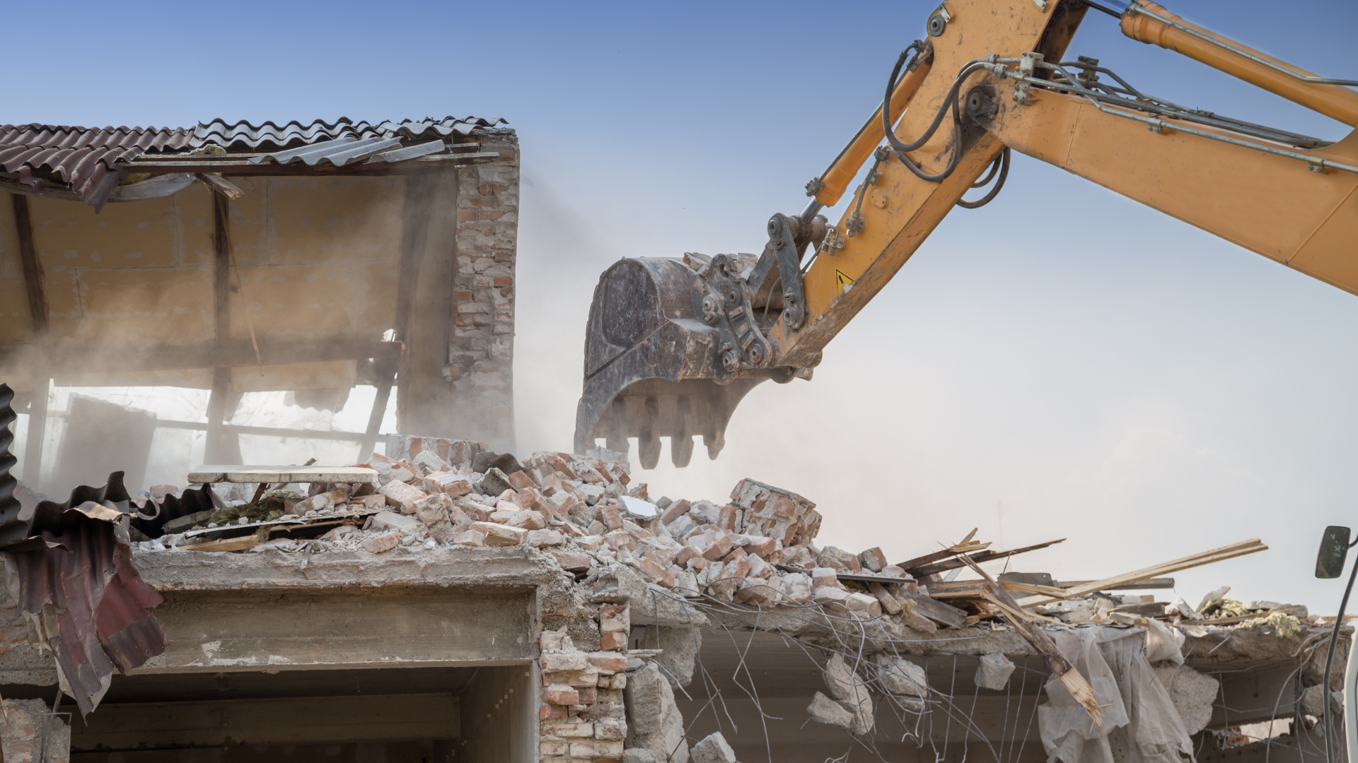 Excavator demolishing a damaged building, with debris falling from the structure.