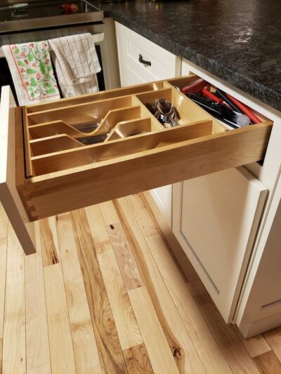Open kitchen drawer with wooden utensil organizer; light wood, black countertop, white cabinets, hardwood floor.