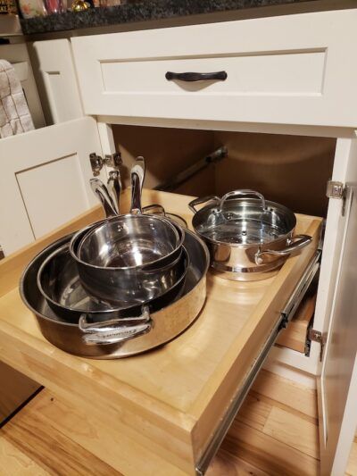Pull-out cabinet drawer with stainless steel pots and pans inside a white kitchen.