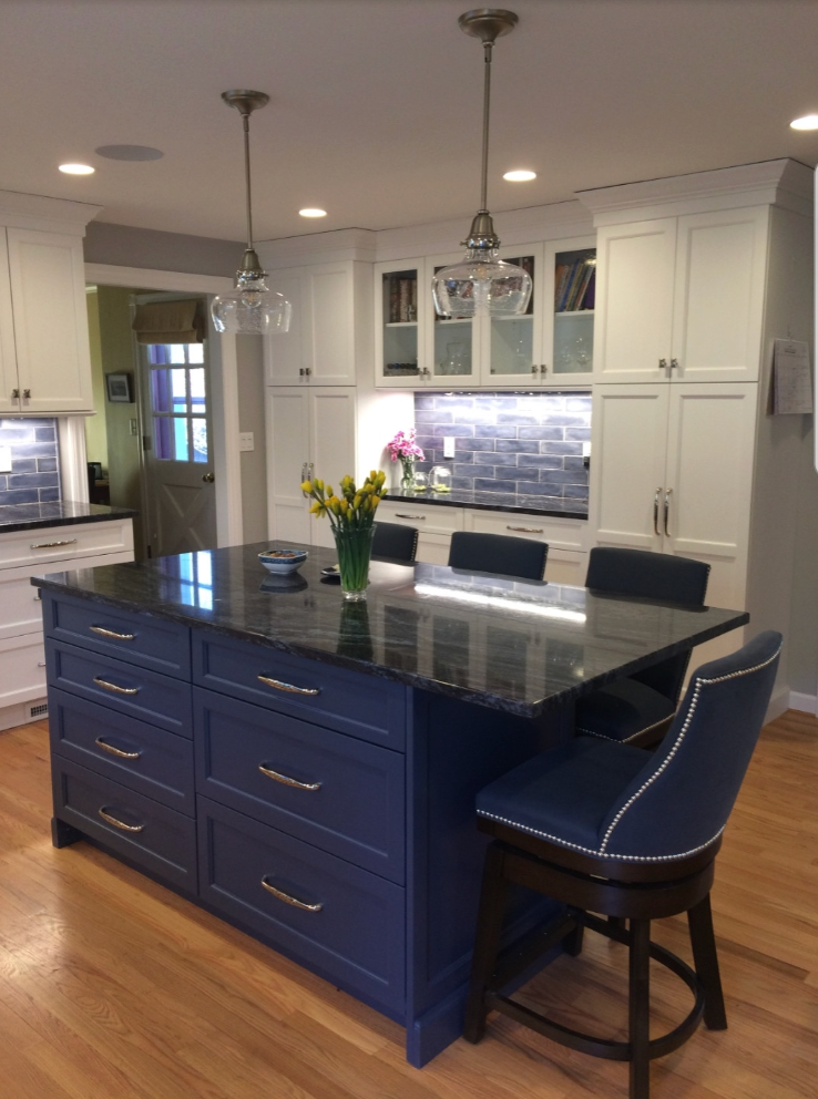 Blue kitchen island with black countertop, drawers, and bar stools; white cabinets, wood floor, and hanging lights.