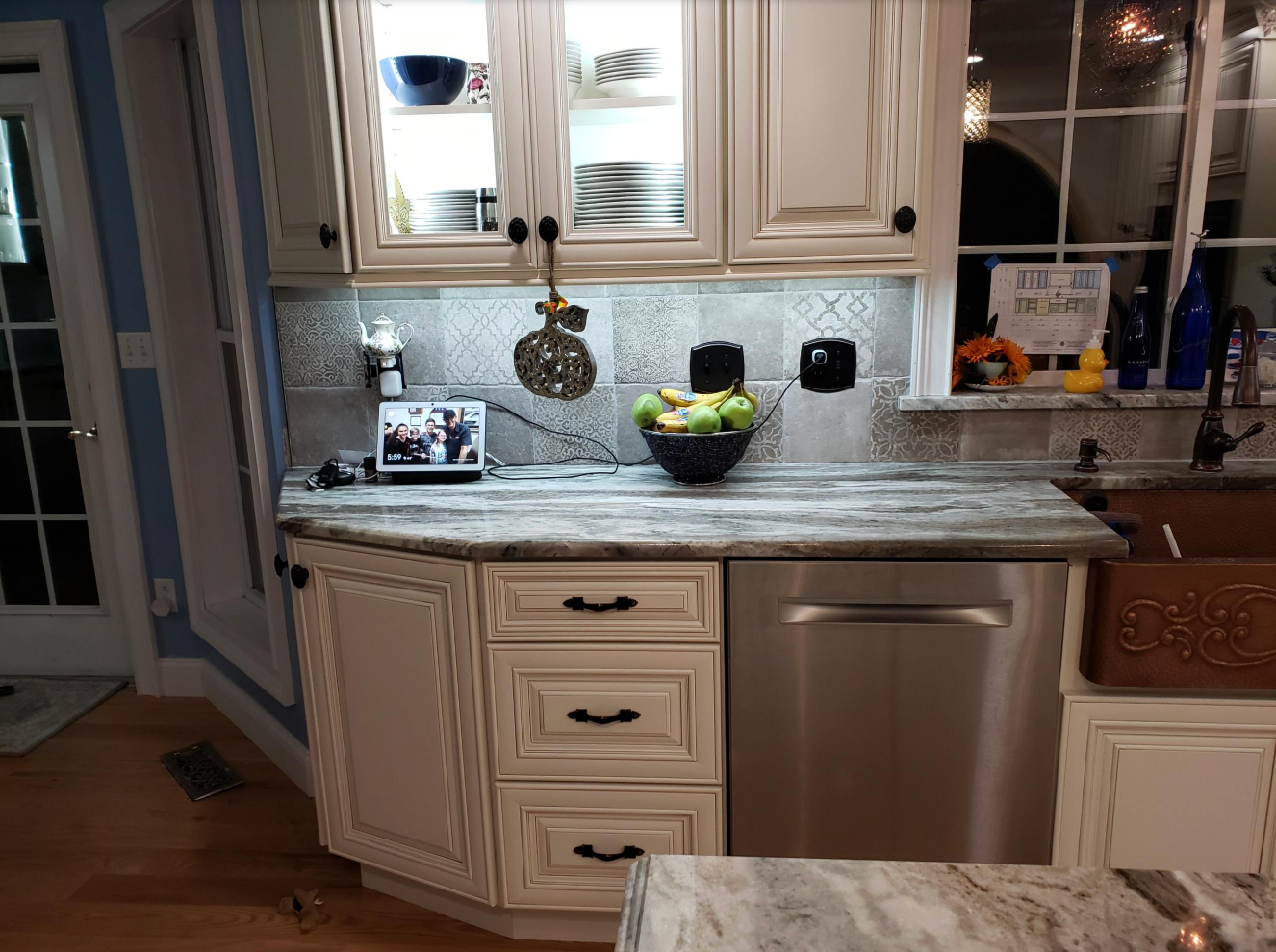Cream-colored kitchen with cabinets, granite countertop, and stainless steel dishwasher. Fruit bowl, utensils, and lit cabinets above.