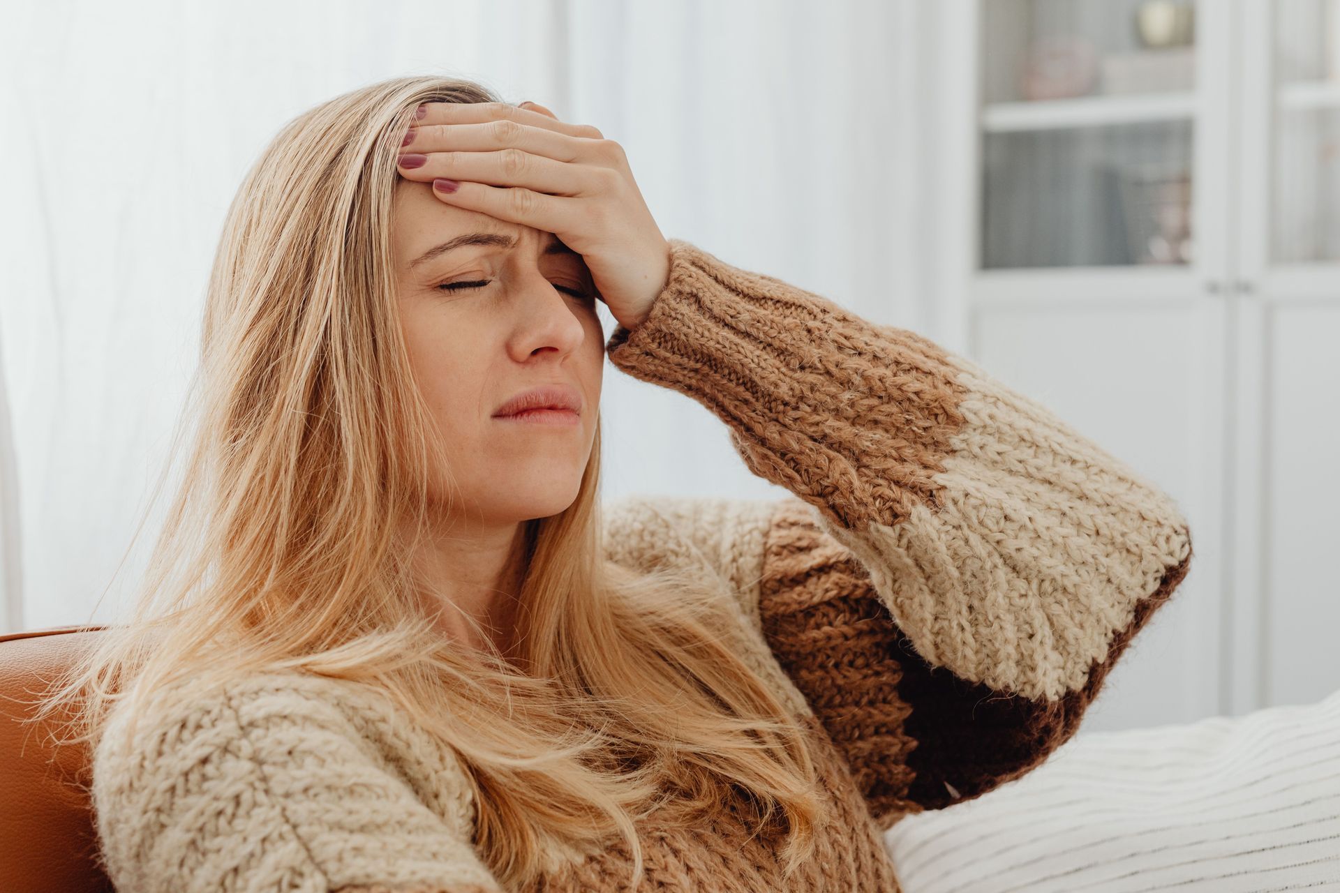 Woman in a beige sweater touching her forehead, looking unwell on a couch indoors