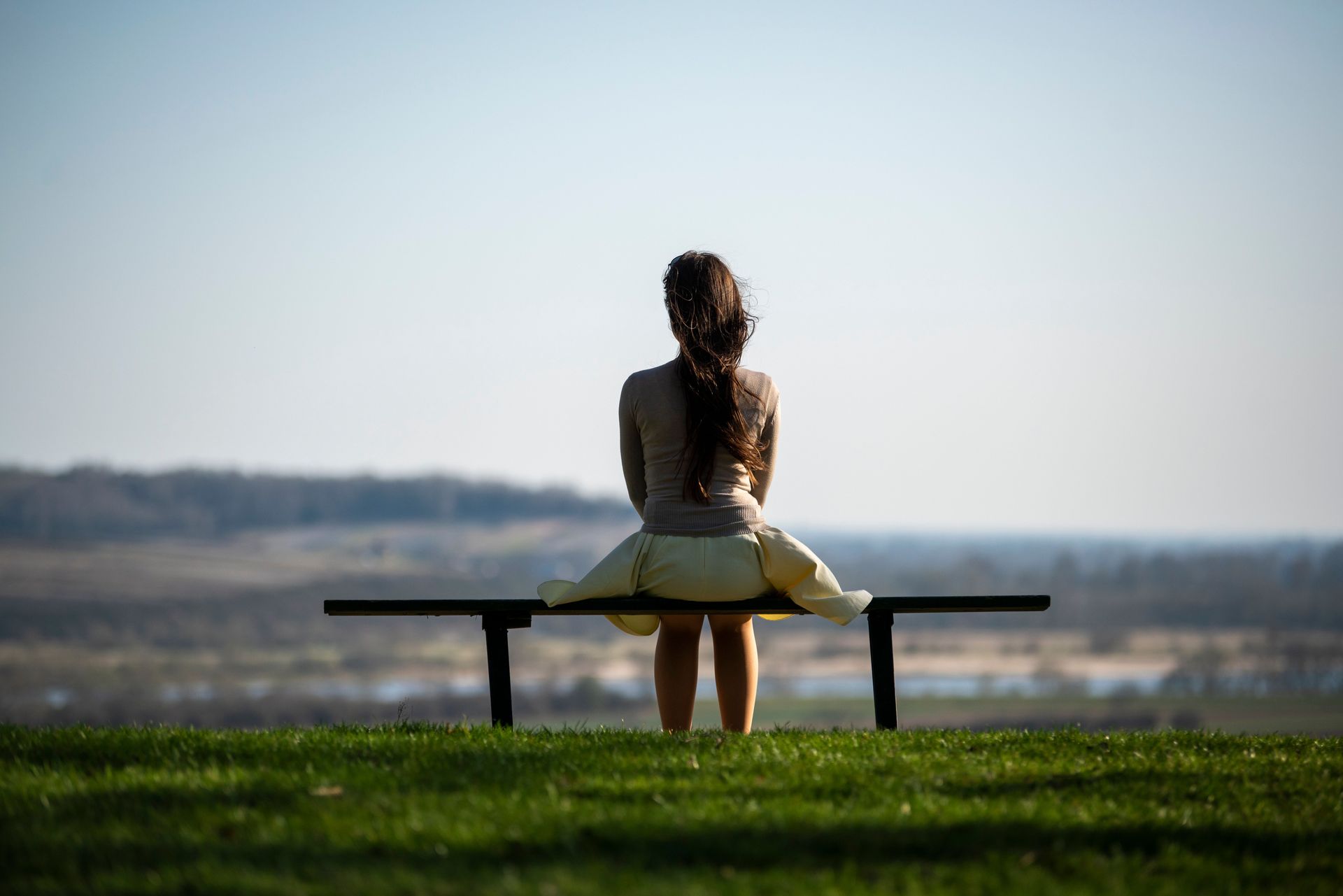 Person sitting on a bench overlooking a lake, with green grass and a hazy horizon.