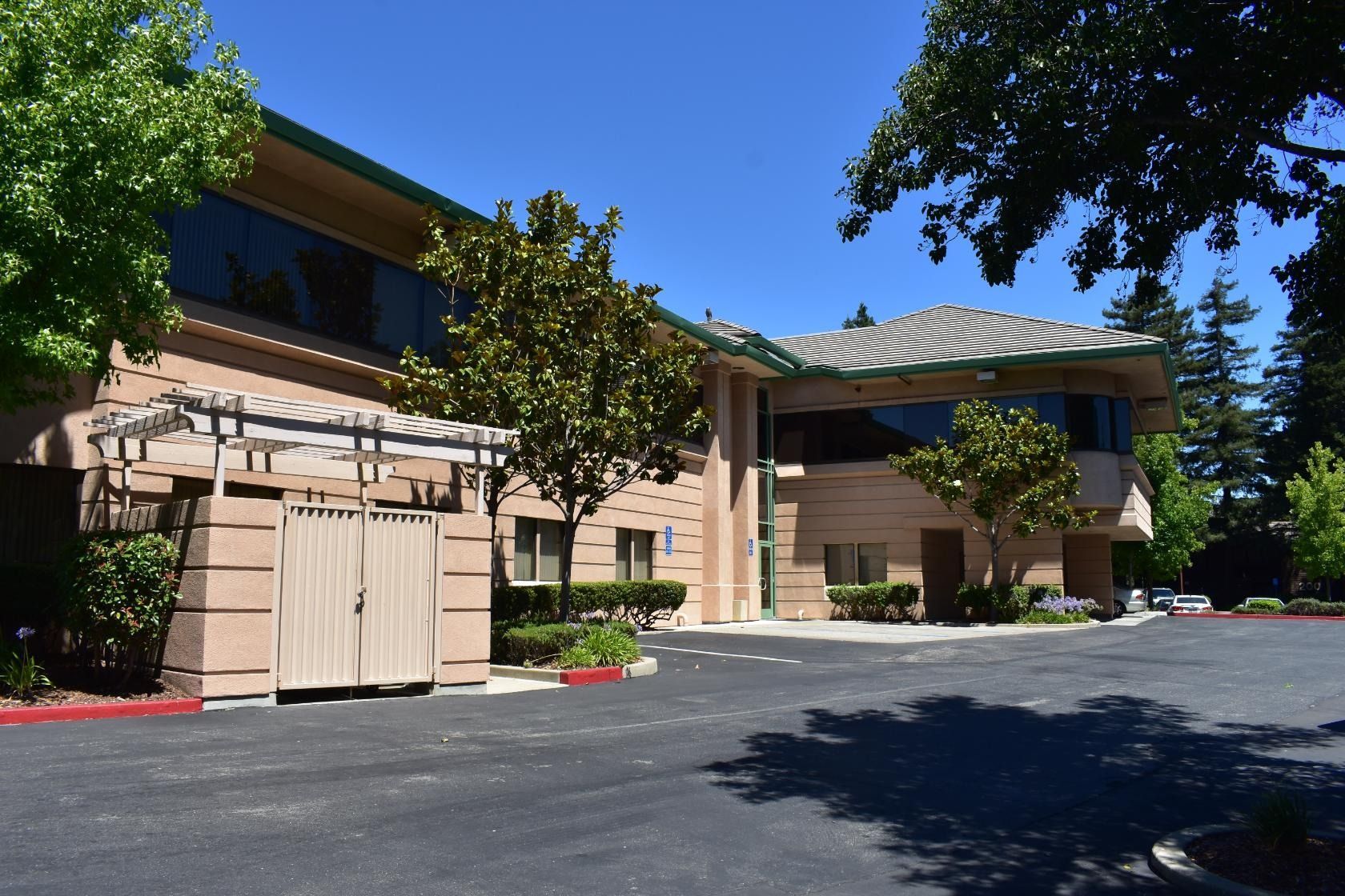 Beige building with large windows and a tiled roof, surrounded by trees and a black asphalt parking lot.