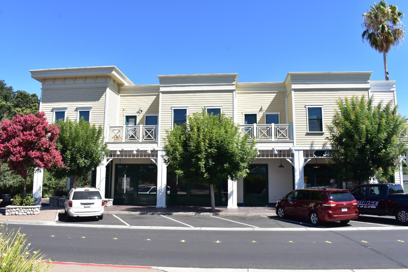 Two-story building with shops, trees, and parked cars under a blue sky.