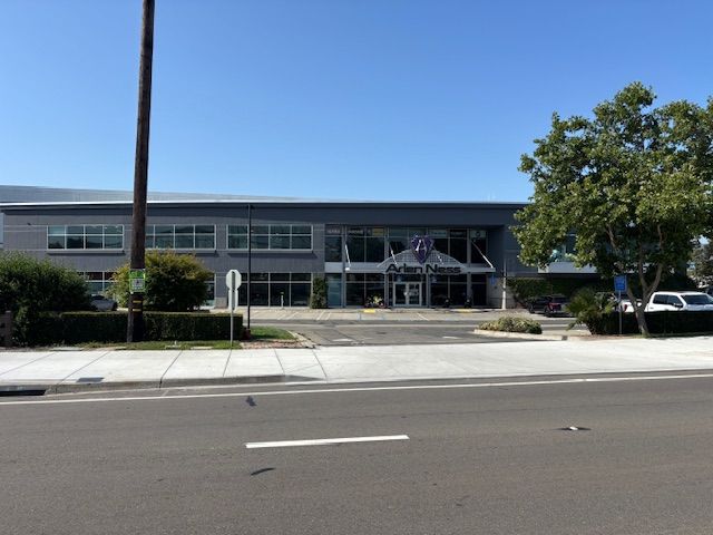 Gray commercial building with glass entrance, on a sunny day. Road in foreground, trees and bushes nearby.