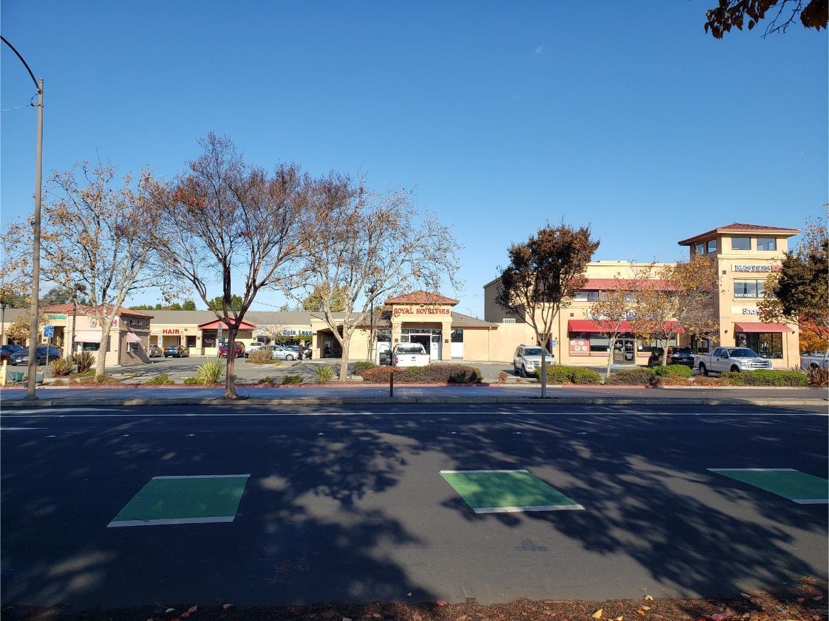 Shops and businesses line a street under a clear blue sky. Trees with autumn leaves and parking spaces are in view.
