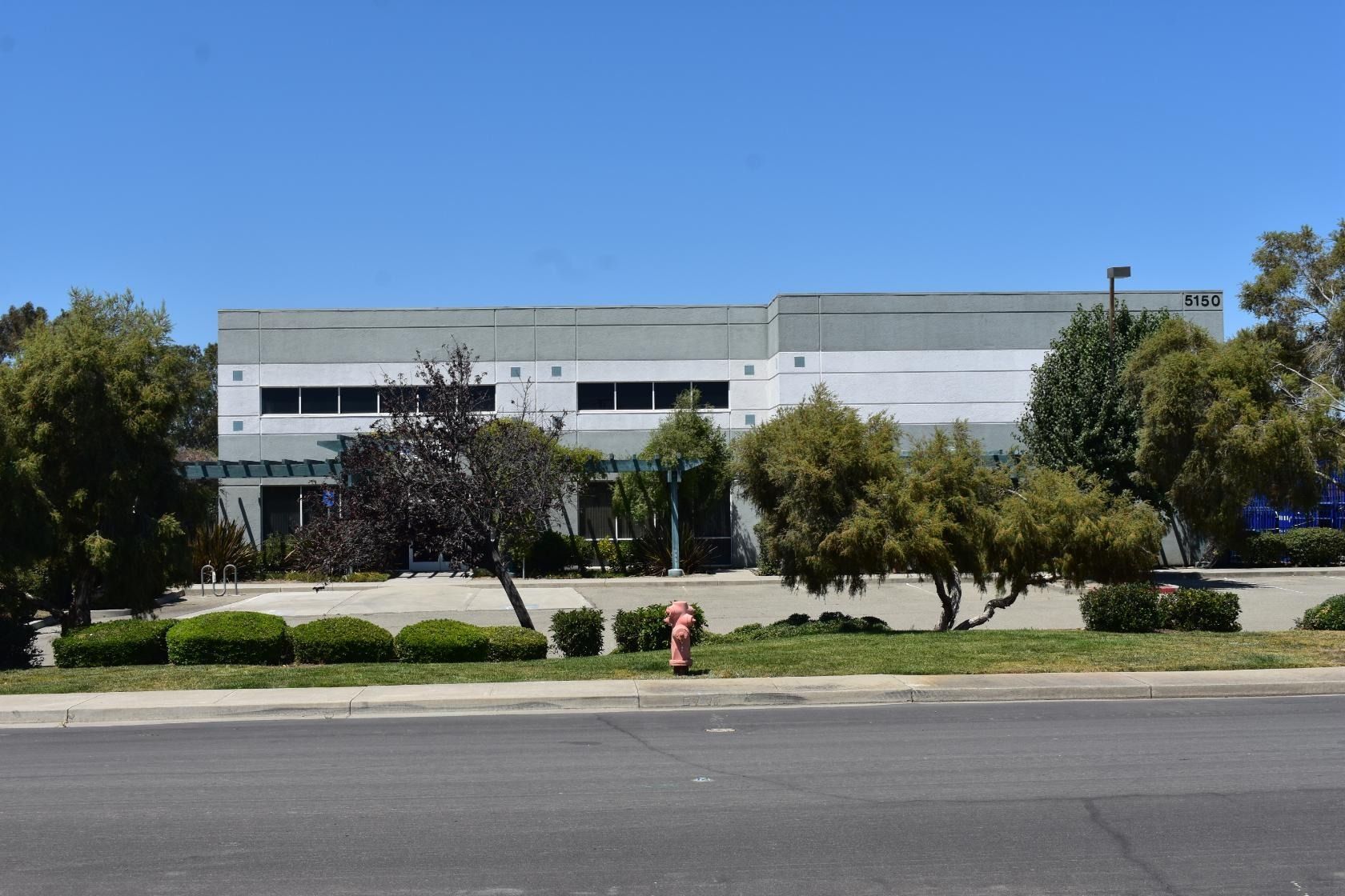 Gray commercial building with windows, trees, a fire hydrant, and a blue sky.
