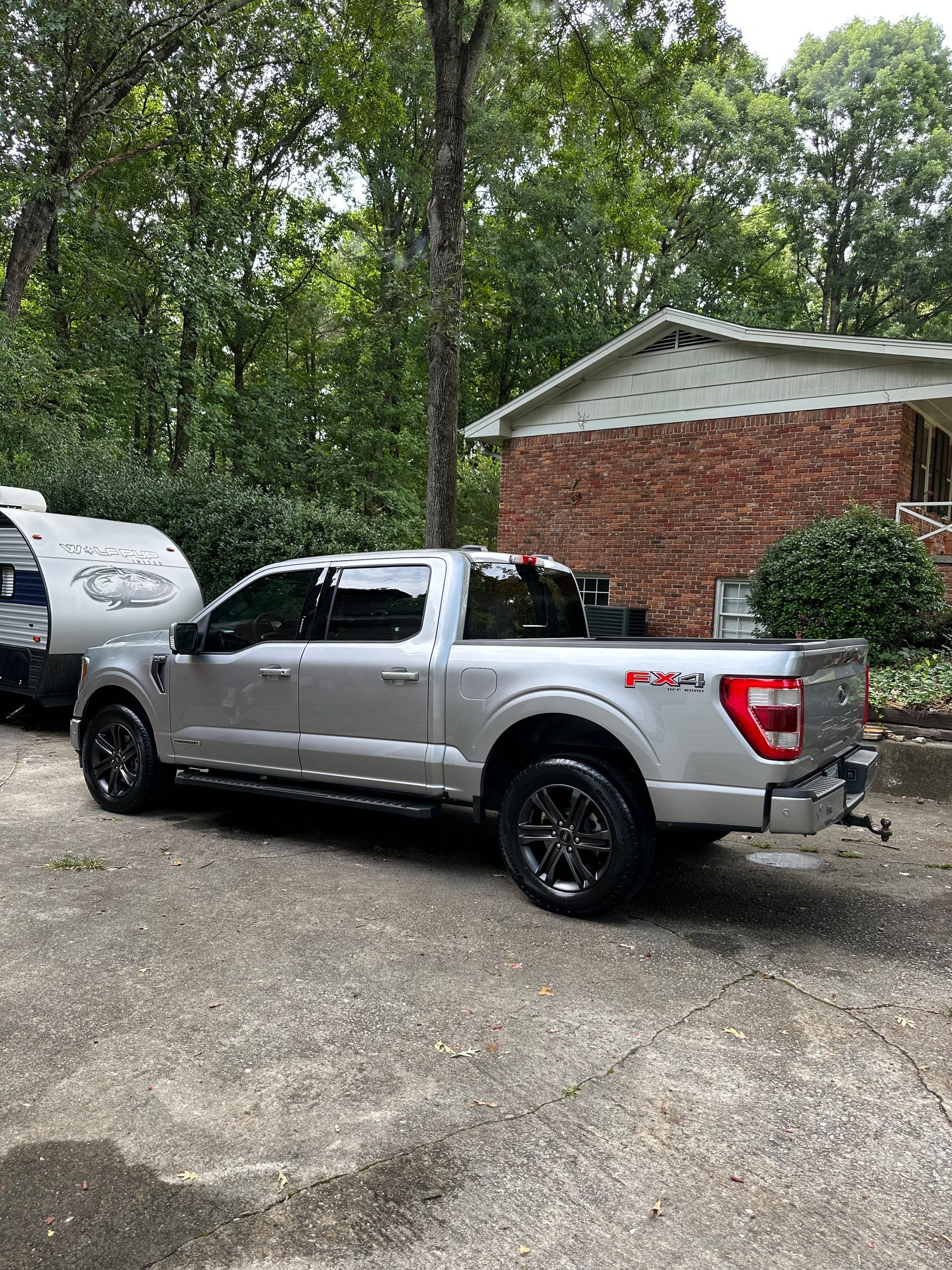 Silver Ford truck towing a camper on a driveway near a brick house and trees.